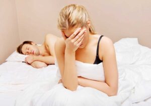 Woman facepalming at the end of her bed while her husband is sleeping.