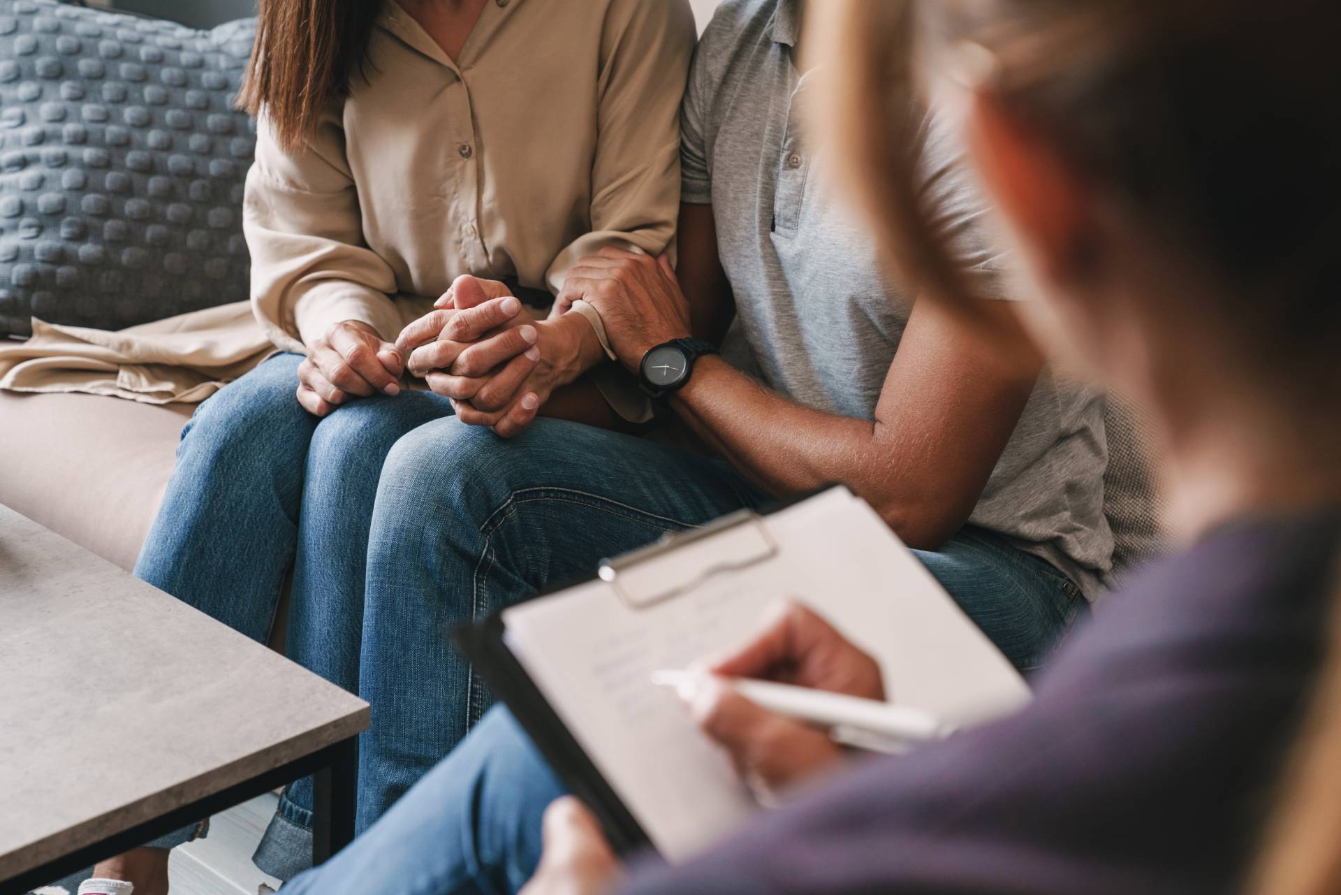 Close-up of a couple holding hands and sitting close together while speaking with a therapist, who is writing notes on a clipboard during a PTSD couples therapy session.