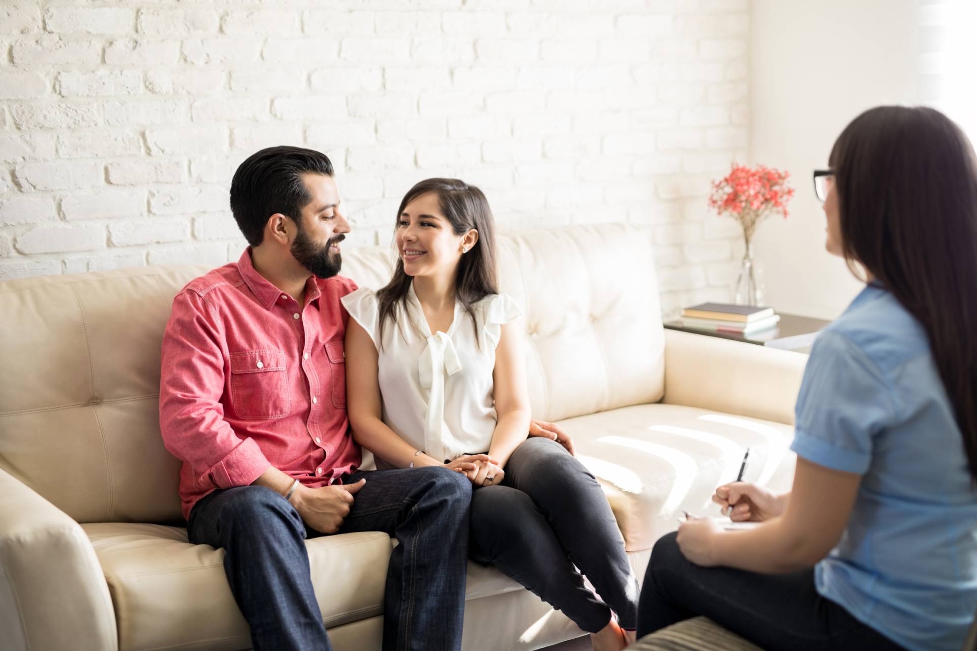 A couple sitting on a beige couch, smiling and holding hands, while speaking with a therapist who is taking notes during a couples therapy session in a bright room with a white brick wall