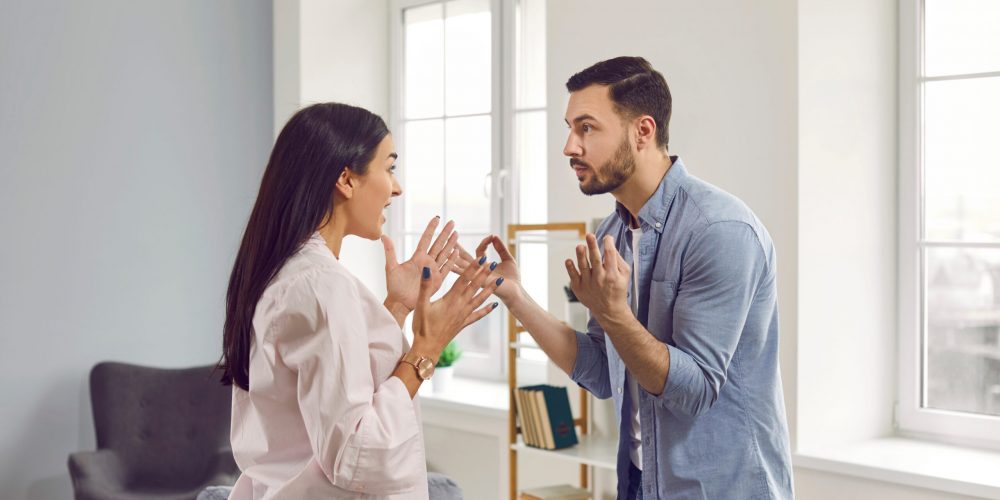 Angry young couple standing at home and arguing in the living room shouting on each other. Frustrated unhappy husband and wife quarreling. Bad marriage,relationships, quarrel and divorce concept.